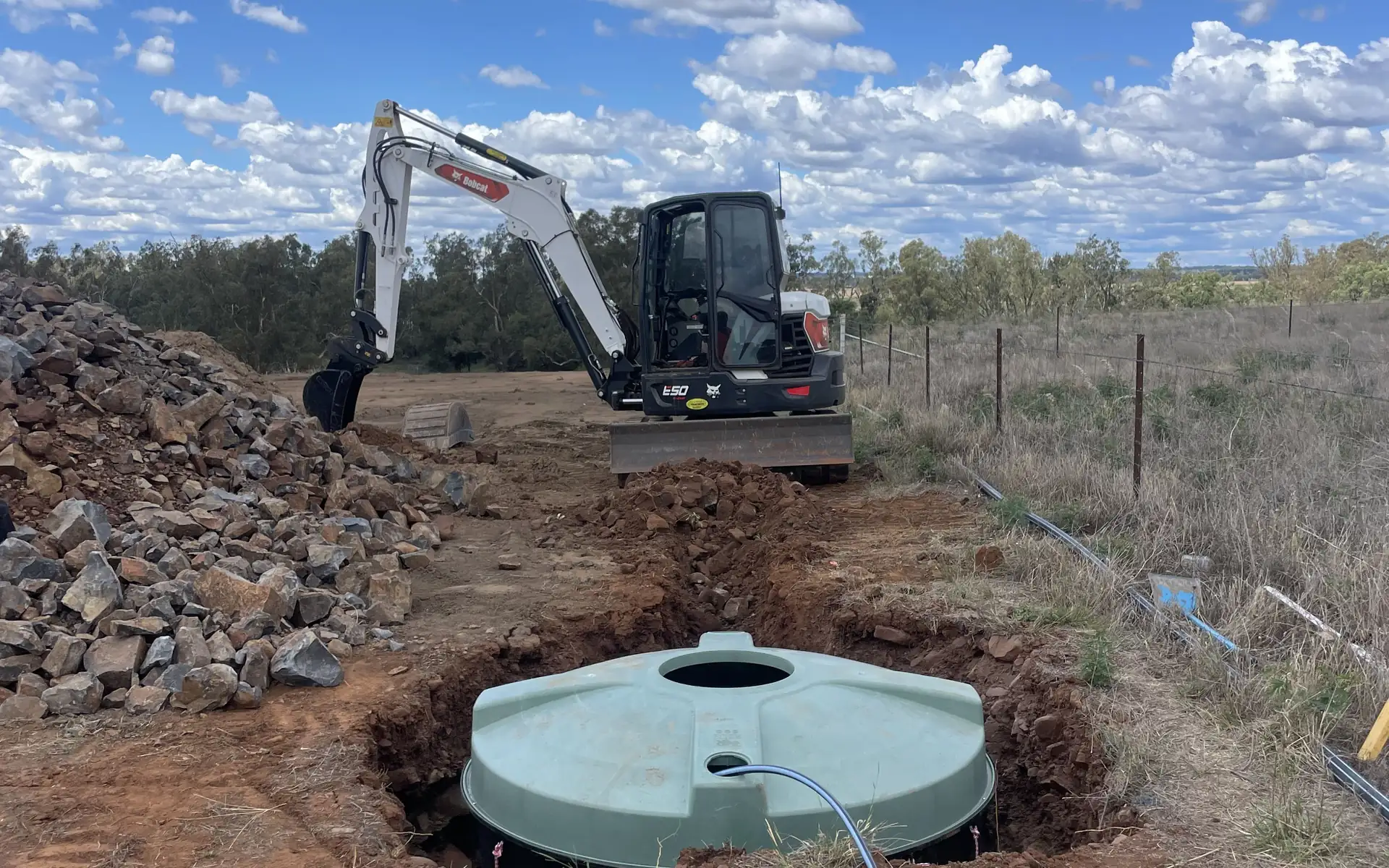 Excavator working on a rural site with an open trench and tank